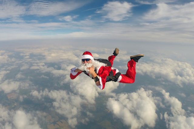 A skydiver in free fall wearing a red santa suit and white beard and sunglasses.